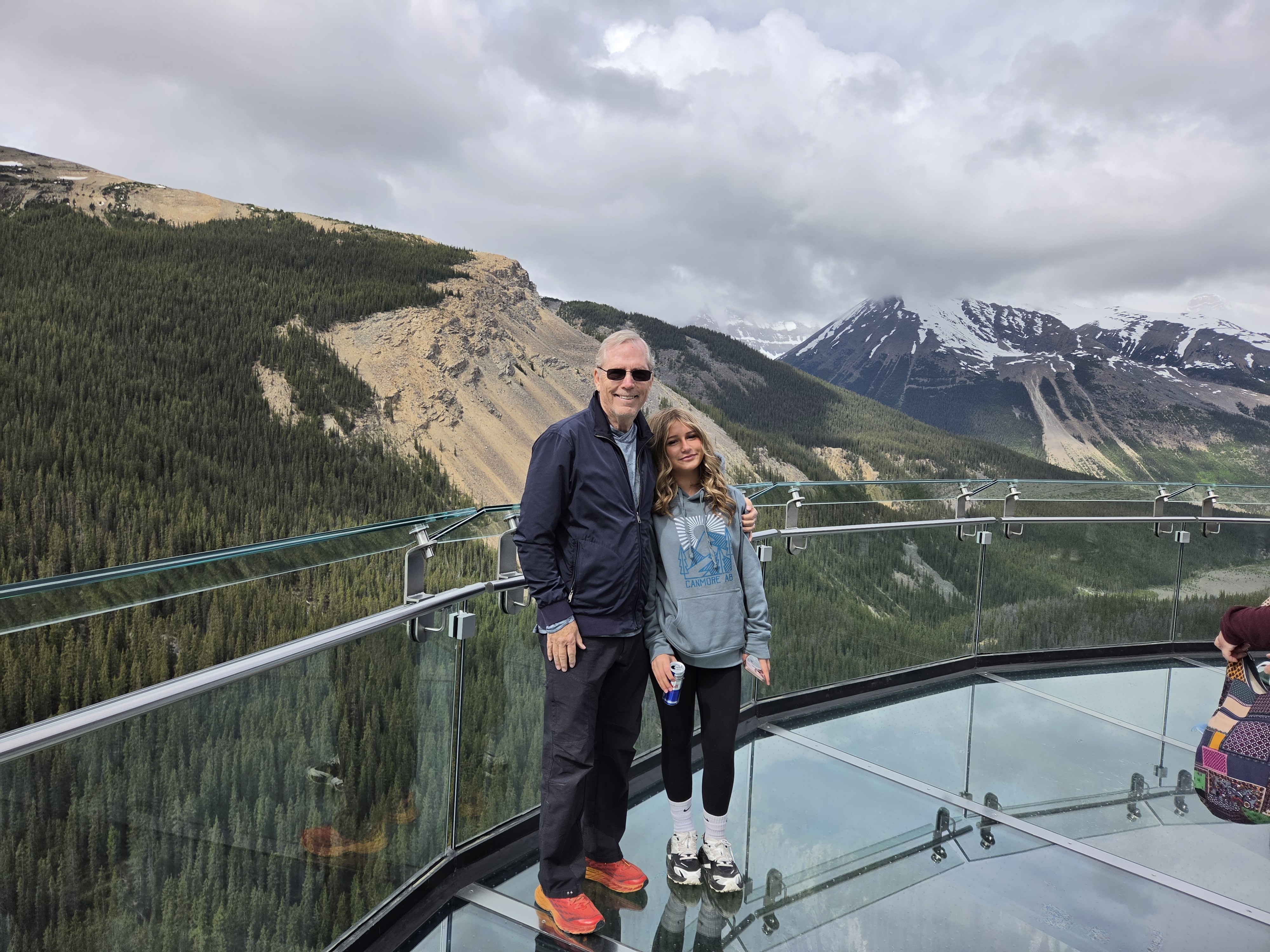 Two people standing on the glass platform of the Skywalk, valley and mountains behind them, the glass floor visible underfoot