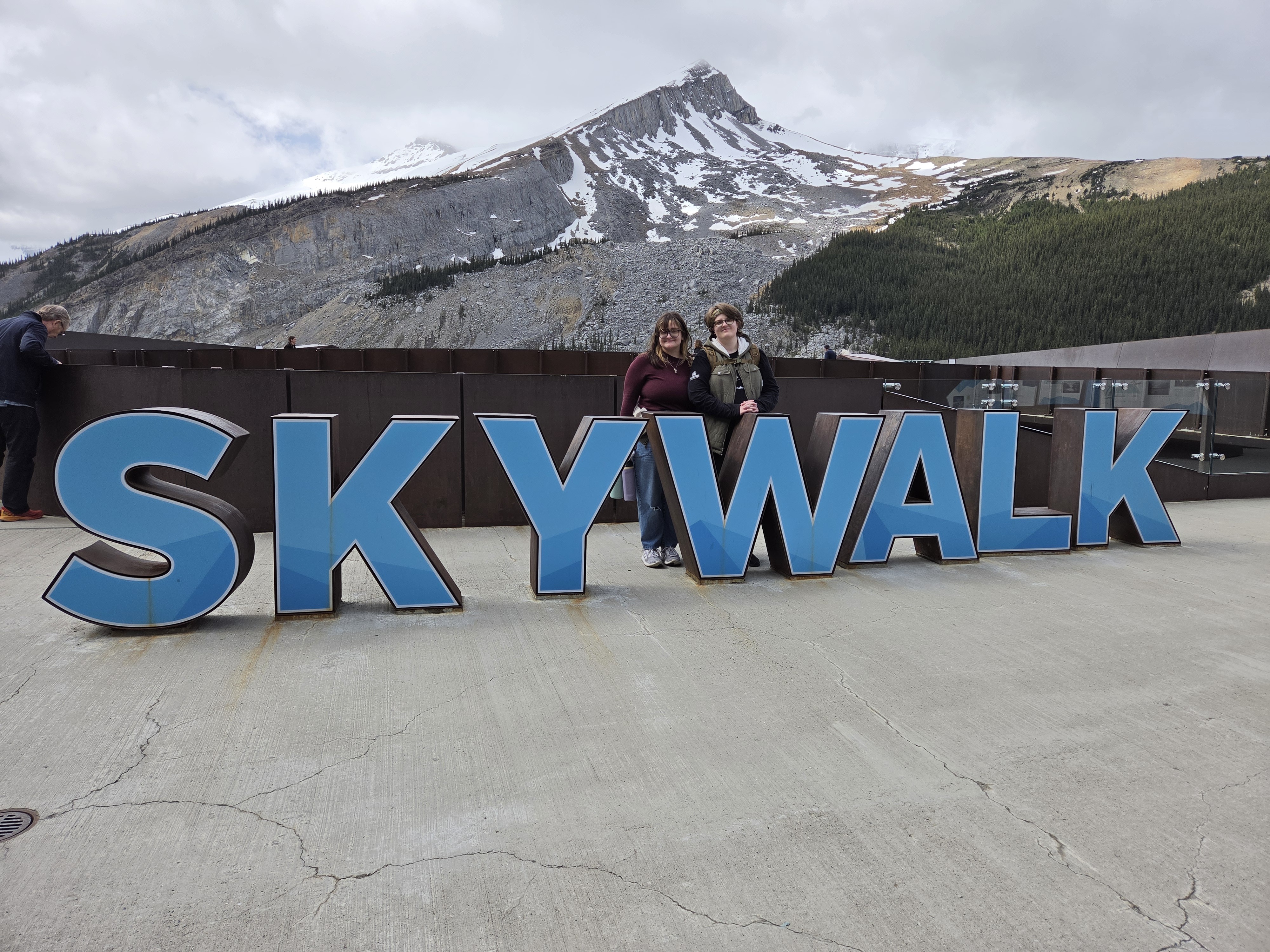 People posing at the large blue “SKYWALK” letters at the end of the walkway, snow-streaked mountains behind