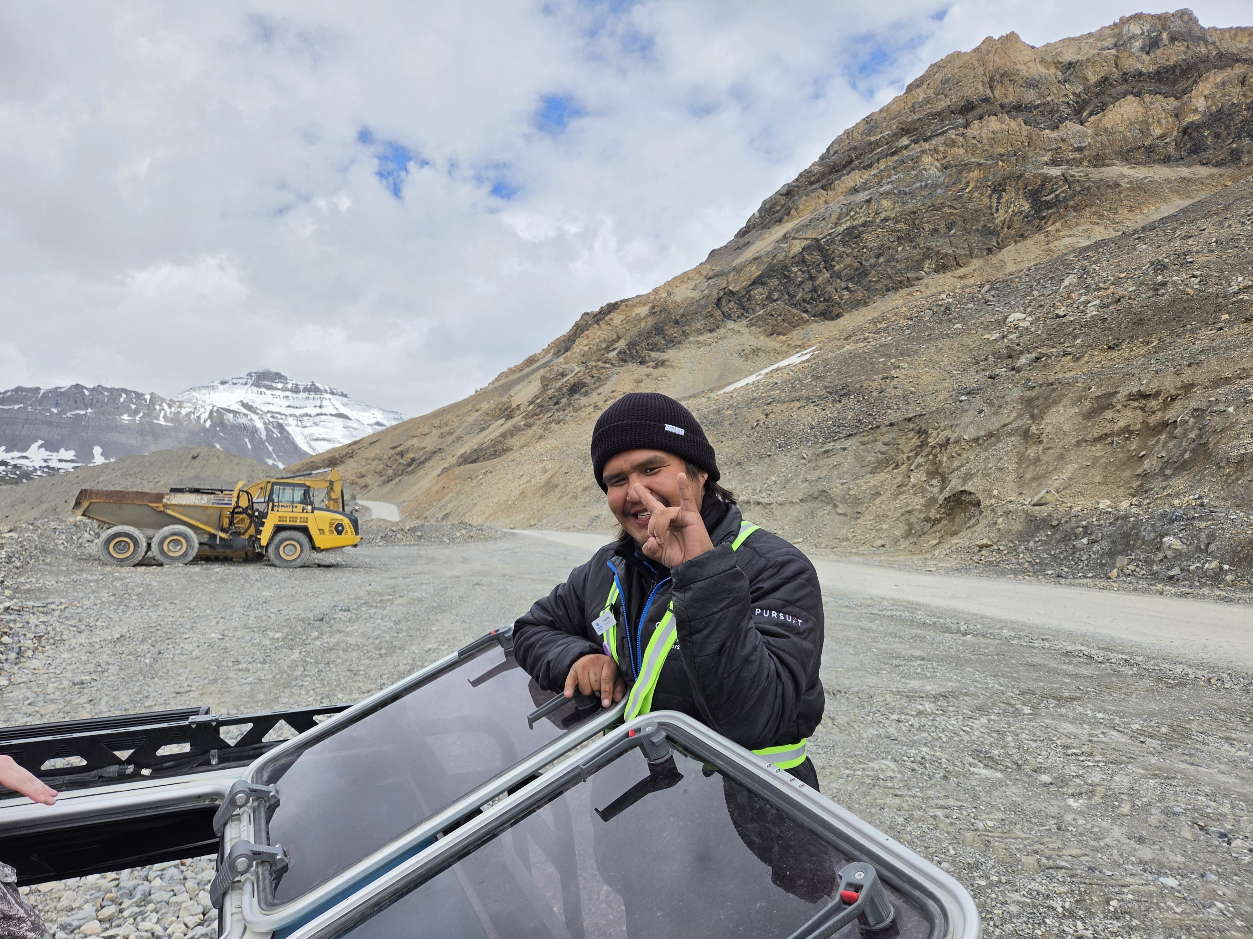 A specialized glacier vehicle with oversized tires on the gravel approach road, a passenger looking out, grey mountains beyond
