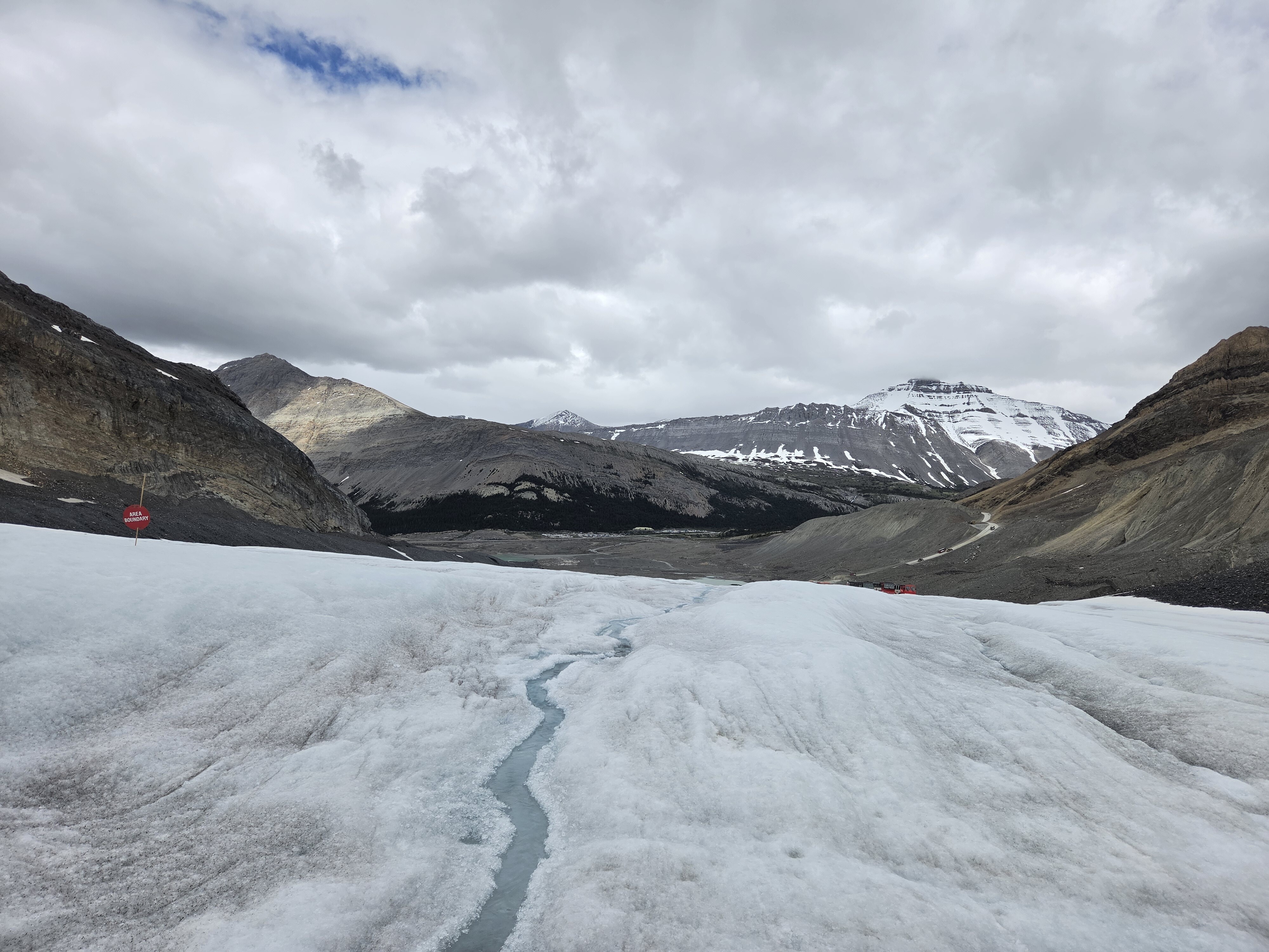 A wide view across the flat grey surface of the Athabasca Glacier, mountains rising on either side under a heavy sky