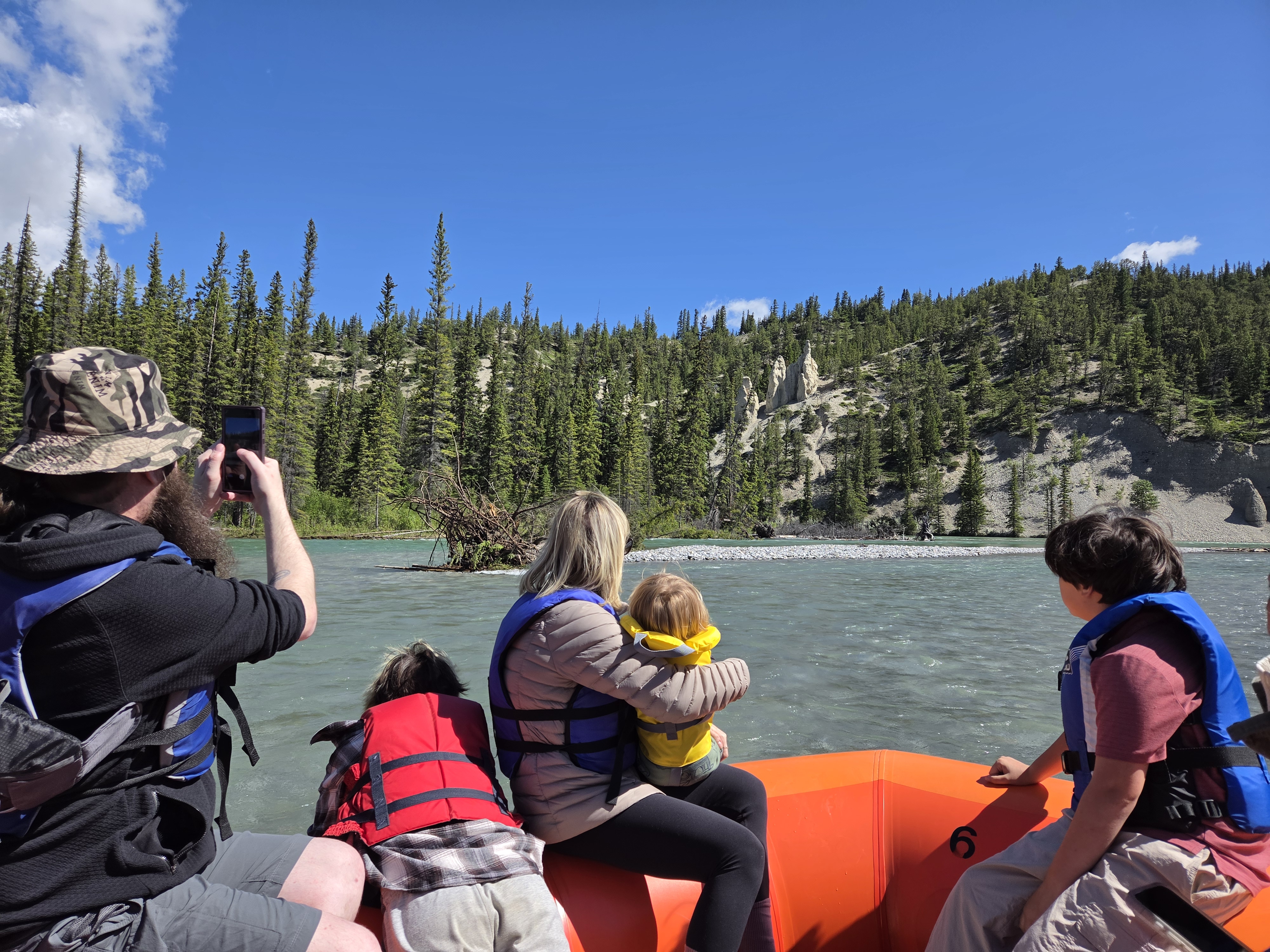 A first-person view down the river from inside the raft, paddlers in orange vests, mountains framing the channel ahead