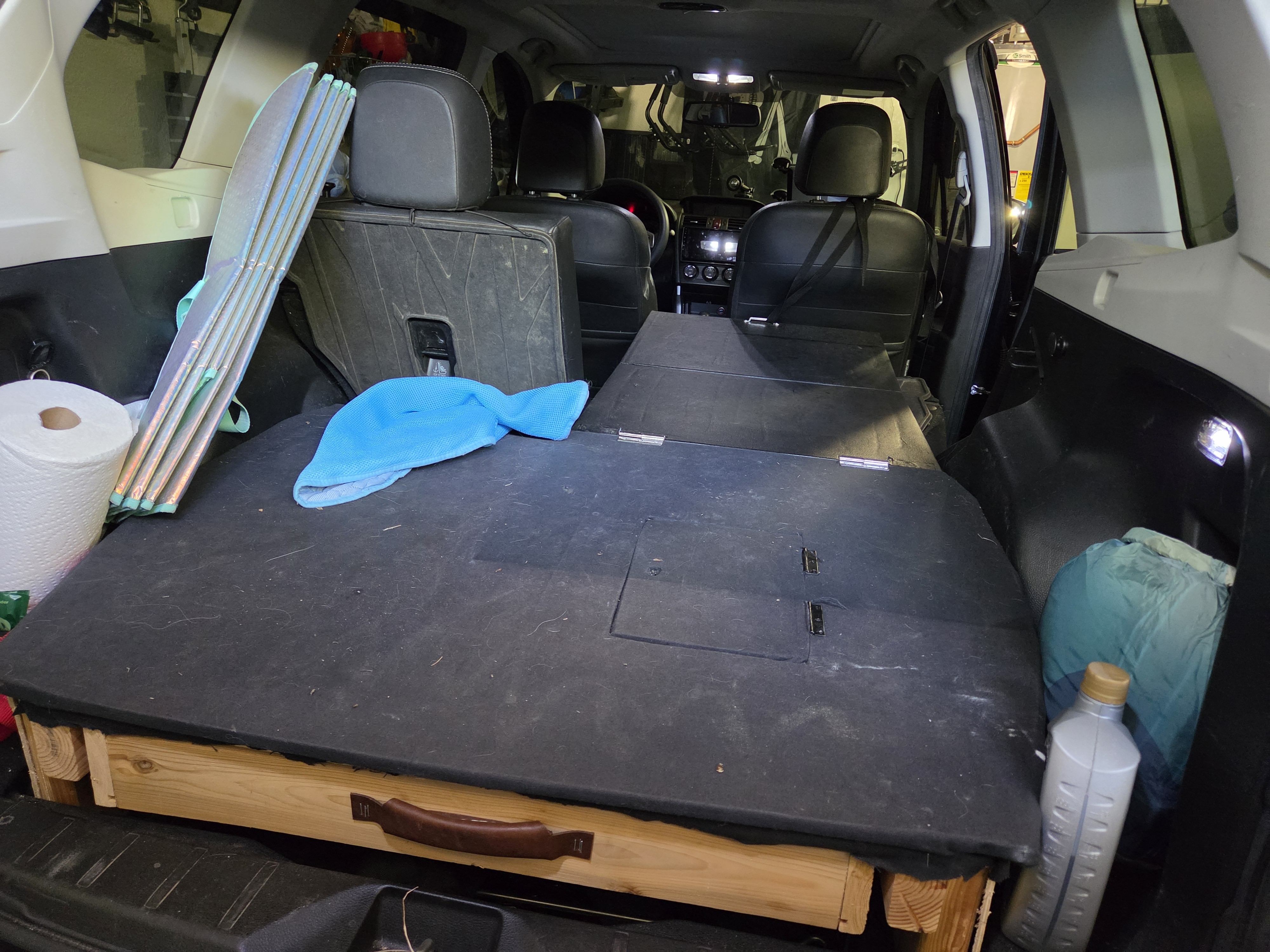 Cargo area with a plywood sleeping platform on a wooden frame, foam topper, folding table, sleeping bag, and water jug