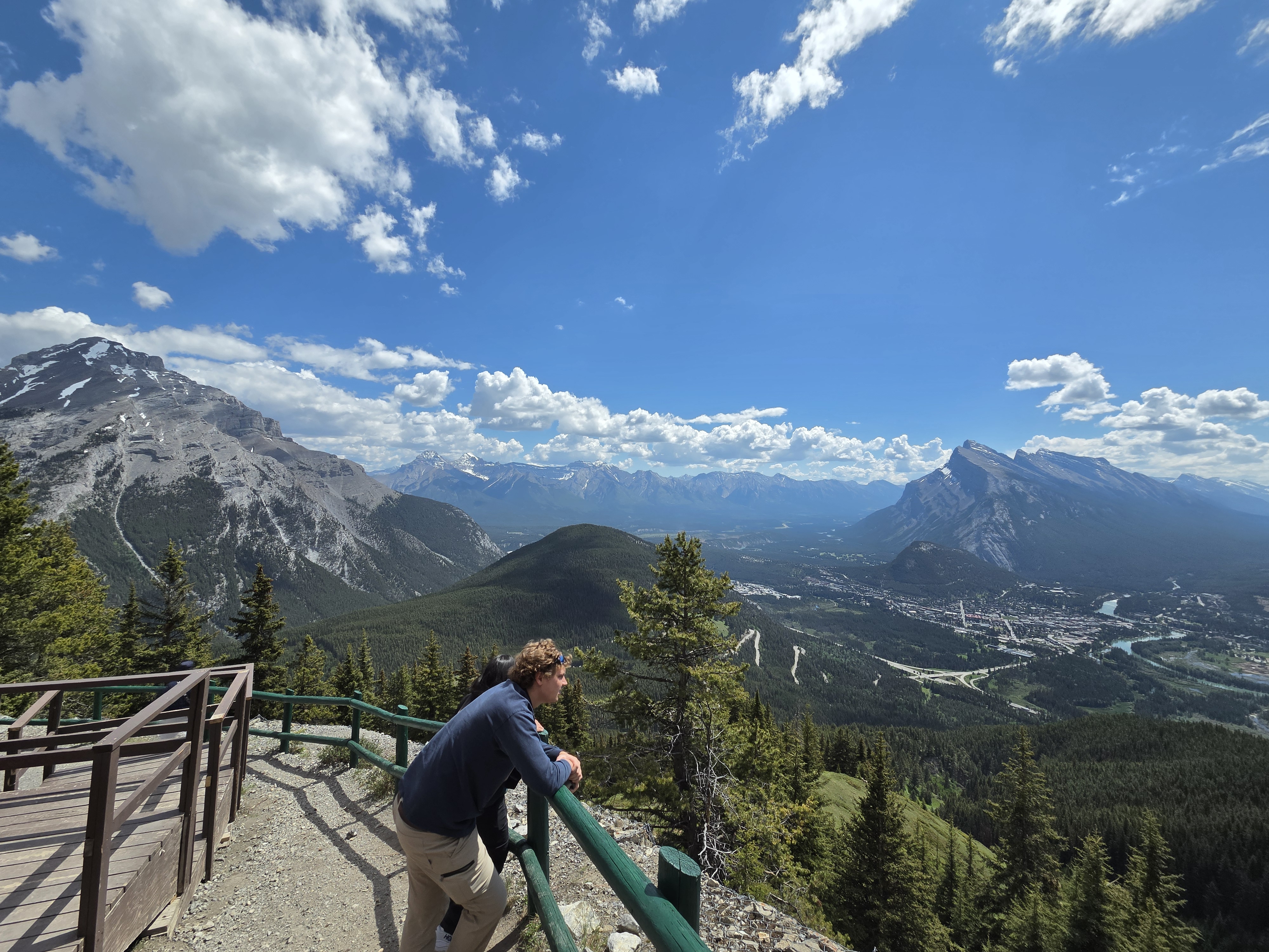 A person standing at a railing on a viewpoint boardwalk, looking out over the valley and distant peaks