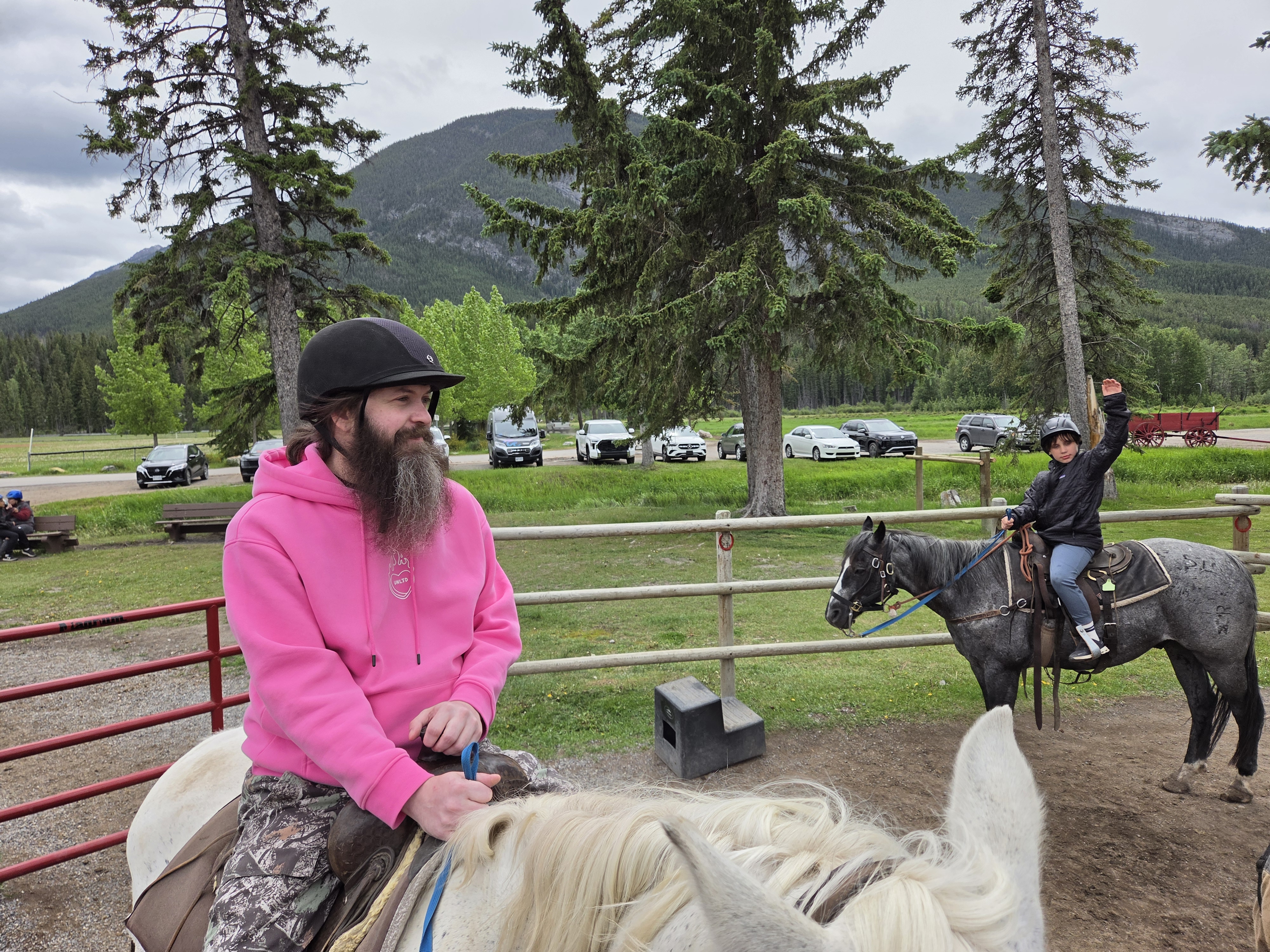 A rider in a pink jacket on horseback at the ranch corral, ready to head out, forest and mountains behind
